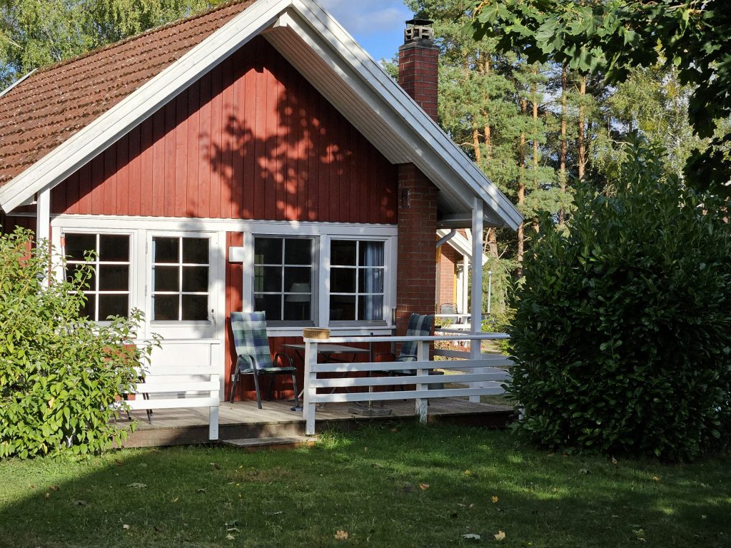 Rotes Ferienhaus Kormoran am Useriner See mit Terrasse, dahinter Bäume. 