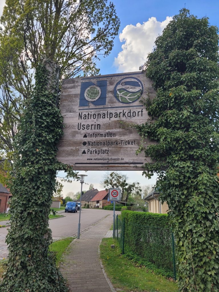 Bild zeigt ein großes Holzschild mit der Beschriftung "Nationalparkdorf Userin - Information - Nationalpark-Ticket - Parkplatz".