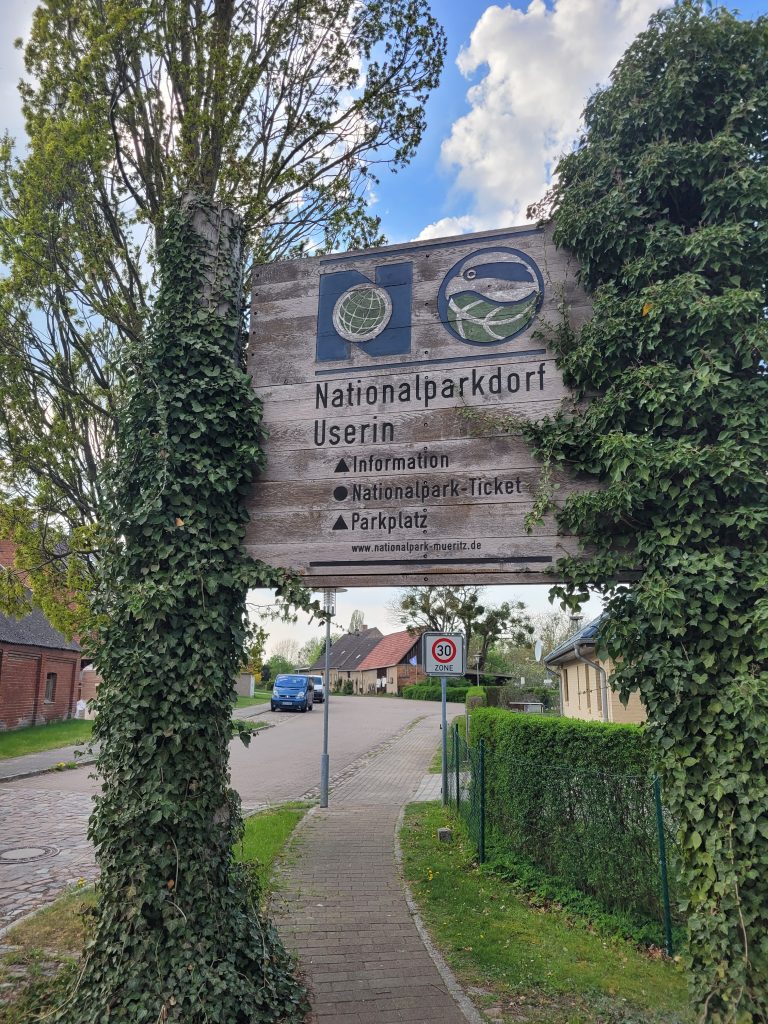 Bild zeigt ein großes Holzschild mit der Beschriftung "Nationalparkdorf Userin - Information - Nationalpark-Ticket - Parkplatz".