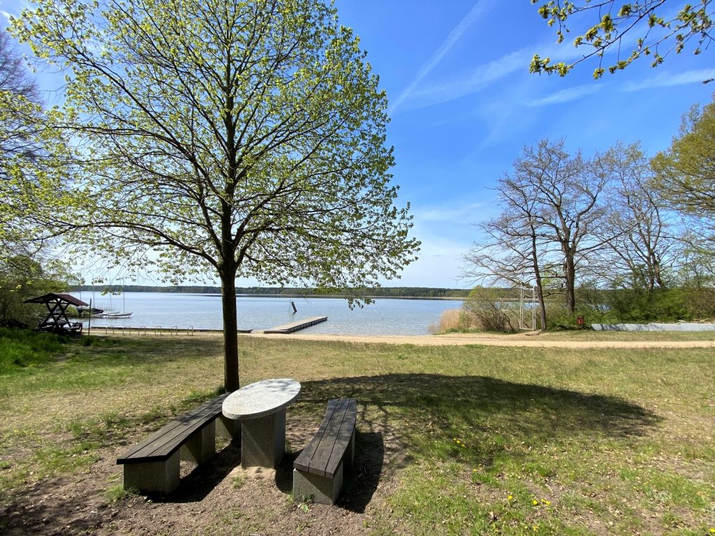 Badestelle in Userin mit Sitzbänken und Tisch, dahinter der See mit Badesteg und Wasserrutsche.