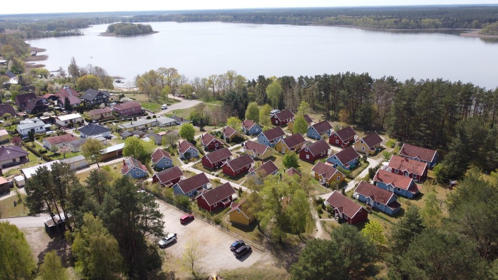 Luftaufnahme zeigt die Ferienhaussiedlung in Userin am Useriner See mit blauen, roten und gelben Holzhäusern im skandinavischen Stil. Im Hintergrund ist der Useriner See und Wald zu sehen.   