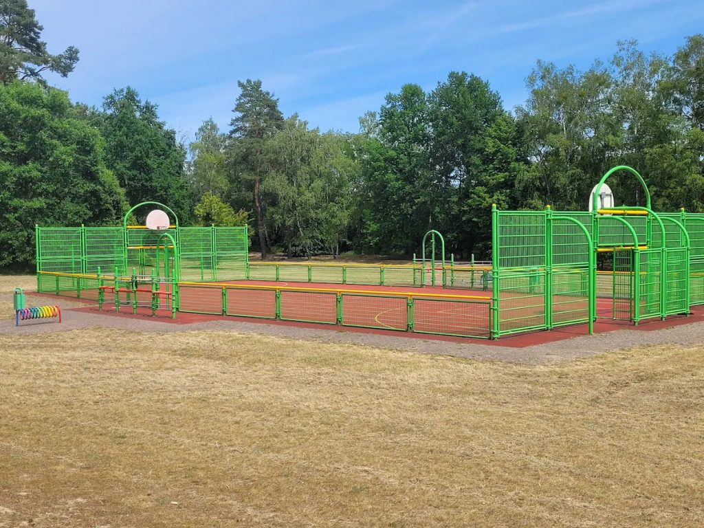 Sportplatz mit Kunststoffboden und Umzäunung, auf dem sich Körbe für Basketball und Tore für Fußball befinden. 