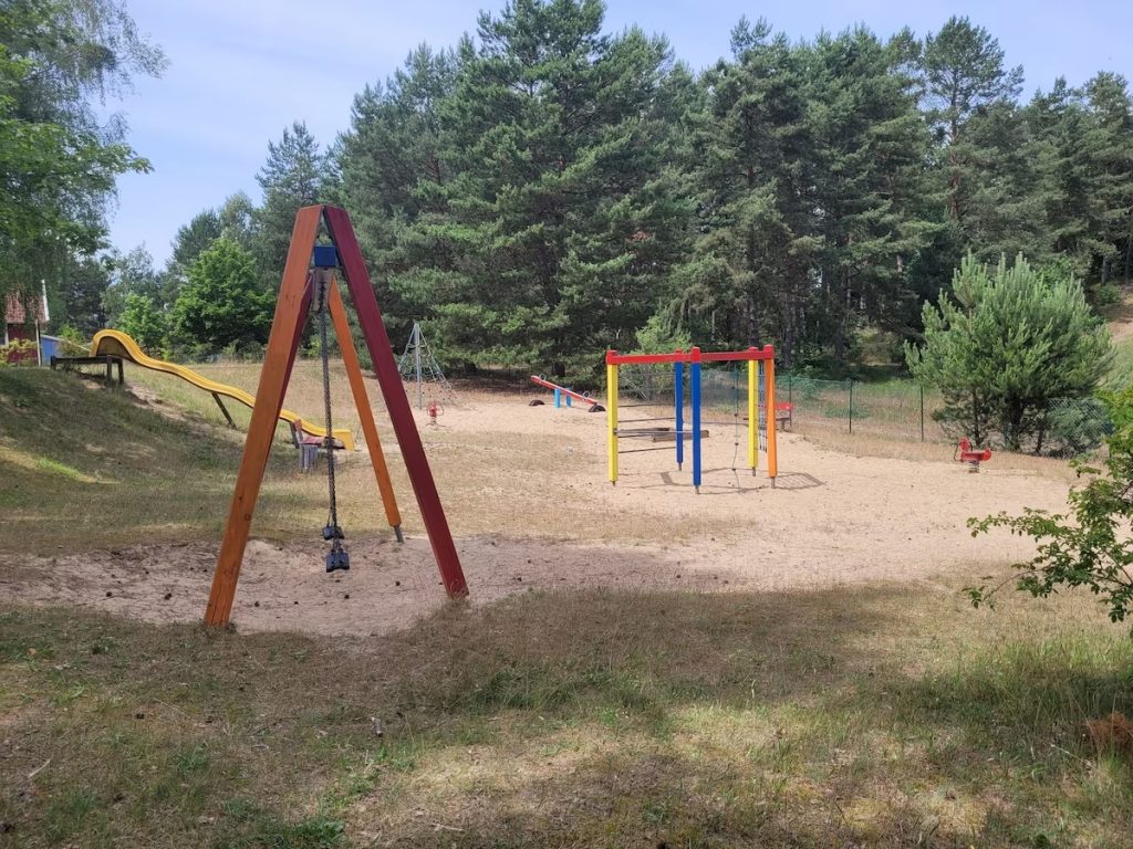 Kinderspielplatz im Ferienzentrum Useriner See mit bunter Schaukel, Rutsche, Klettergerüsten, Wippe und Sand vor grünen Bäumen. 