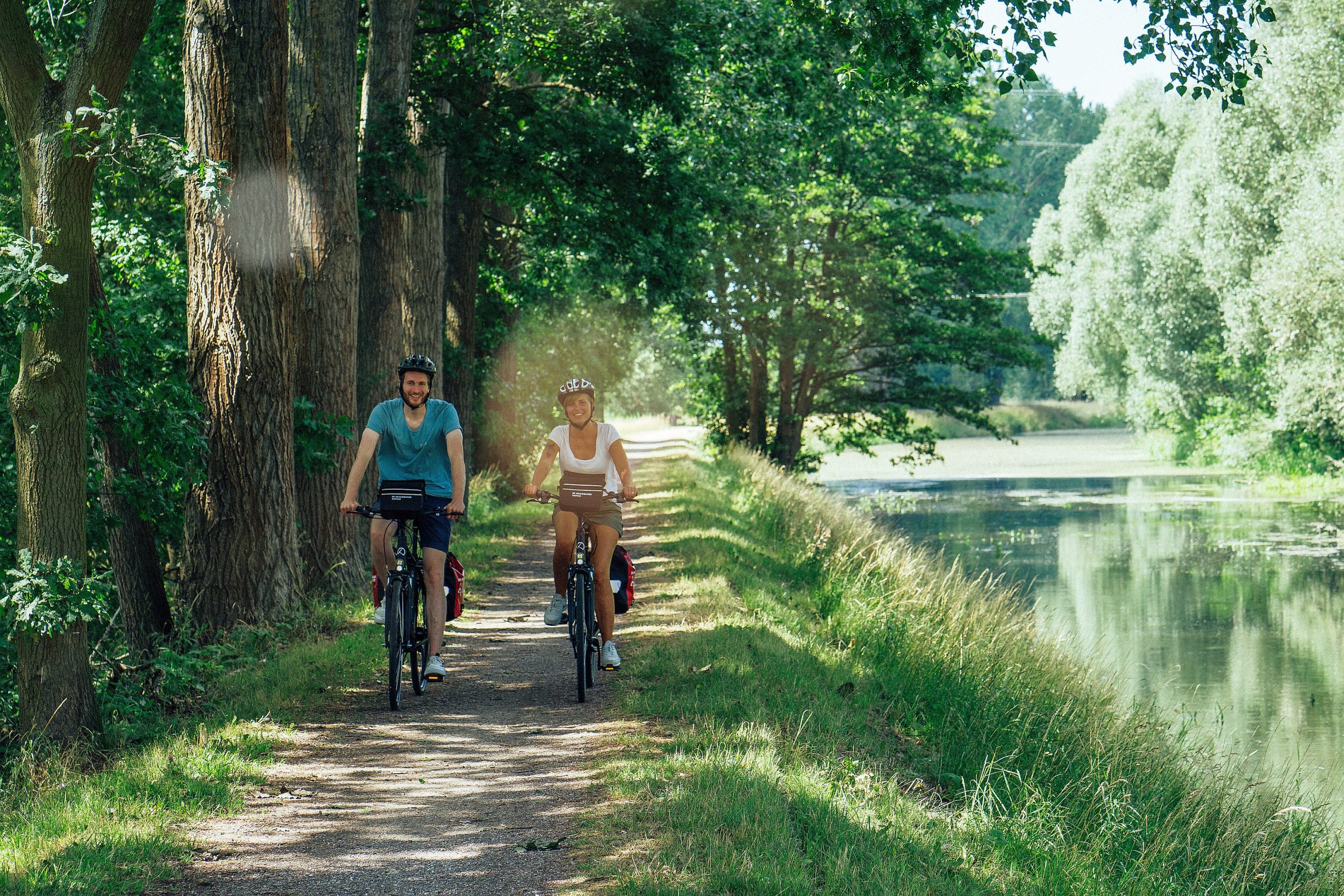 Zwei Radfahrer fahren auf einem Schotterweg, auf der einen Seite hohe Bäume, auf der anderen Seite ein Gewässer.