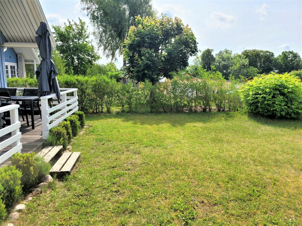 Garten mit Wiese und Hecken und Bäumen, links im Bild der Rand einer Terrasse mit Sonnenschirm und Sitzgruppe